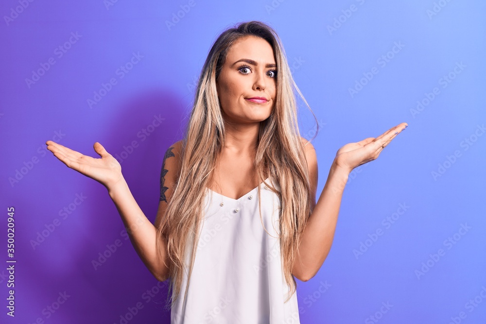 Young beautiful blonde woman wearing white t-shirt standing over isolated purple background clueless and confused with open arms, no idea and doubtful face. Young beautiful blonde woman wearing white t-shirt standing over isolated purple background clueless and confused with open arms, no idea and doubtful face.