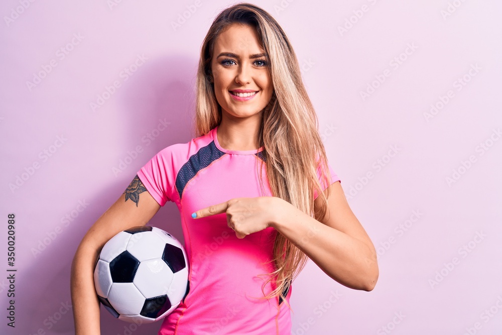 Young beautiful blonde player woman playing soccer holding football ball over pink background smiling happy pointing with hand and finger Young beautiful blonde player woman playing soccer holding football ball over pink background smiling happy pointing with hand and finger