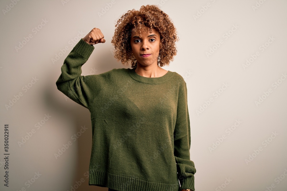 Beautiful african american woman with curly hair wearing casual sweater over white background Strong person showing arm muscle, confident and proud of power Beautiful african american woman with curly hair wearing casual sweater over white background Strong person showing arm muscle, confident and proud of power