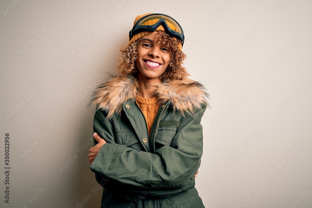 Young african american skier woman with curly hair wearing snow sportswear and ski goggles happy face smiling with crossed arms looking at the camera. Positive person.