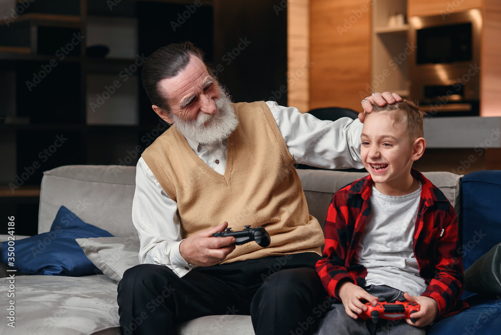 Cute little boy with grandfather sitting on sofa and playing video game with game pad