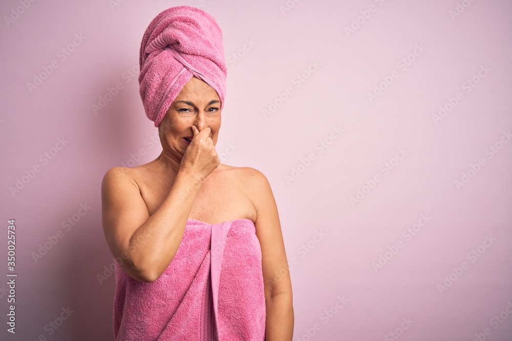 Middle age woman wearing bath towel from beauty body care over pink background smelling something stinky and disgusting, intolerable smell, holding breath with fingers on nose. Bad smell Middle age woman wearing bath towel from beauty body care over pink background smelling something stinky and disgusting, intolerable smell, holding breath with fingers on nose. Bad smell