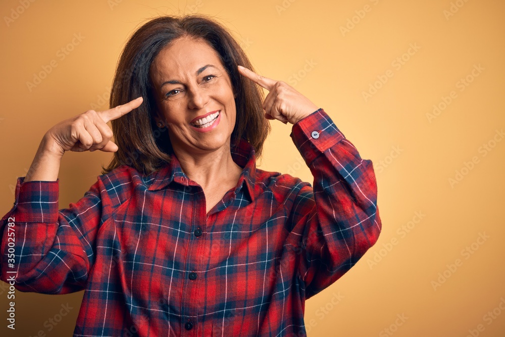 Middle age beautiful woman wearing casual shirt standing over isolated yellow background smiling pointing to head with both hands finger, great idea or thought, good memory Middle age beautiful woman wearing casual shirt standing over isolated yellow background smiling pointing to head with both hands finger, great idea or thought, good memory