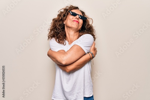 Middle age beautiful woman wearing funny thug life sunglasses over white background hugging oneself happy and positive, smiling confident. Self love and self care