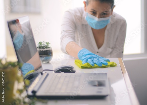 Woman in surgical mask cleaning the desk and laptop with disinfectant and a cloth to disinfect bacteria and viruses. Health and hygiene in the office and at home.