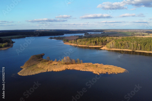 River island and forest aerial view. Storage reservoir. Aerial shot of spring landscape with blue sky and white clouds, flat terrain