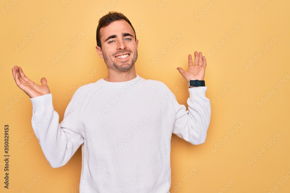 Young handsome man with blue eyes wearing casual sweater standing over yellow background clueless and confused expression with arms and hands raised. Doubt concept. Young handsome man with blue eyes wearing casual sweater standing over yellow background clueless and confused expression with arms and hands raised. Doubt concept.