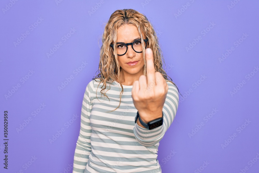 Beautiful blonde woman wearing casual striped t-shirt and glasses over purple background Showing middle finger, impolite and rude fuck off expression