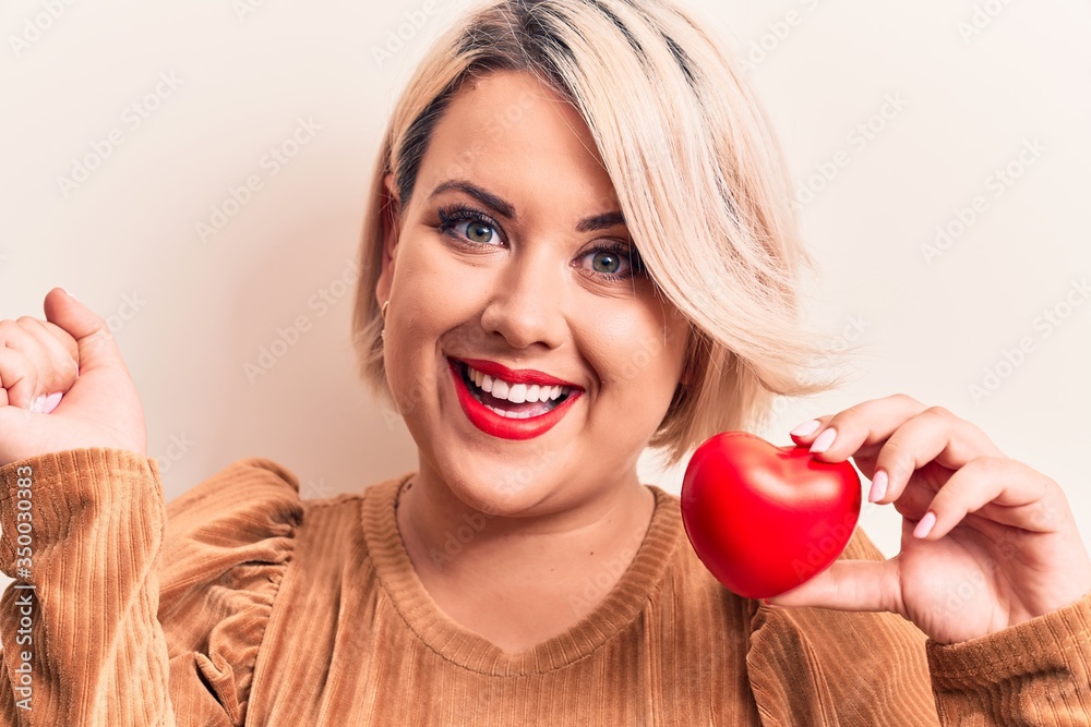 Young beautiful blonde plus size woman holding plastic heart over isolated white background screaming proud, celebrating victory and success very excited with raised arm Young beautiful blonde plus size woman holding plastic heart over isolated white background screaming proud, celebrating victory and success very excited with raised arm