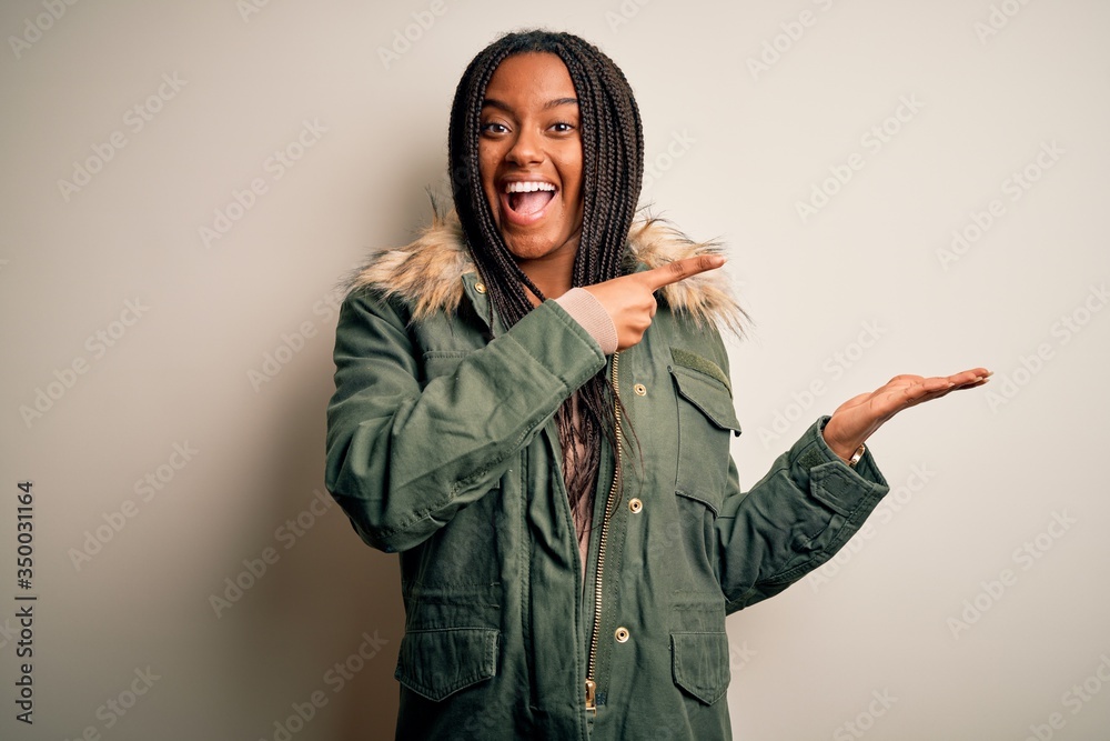 Young african american woman wearing winter parka coat over isolated background amazed and smiling to the camera while presenting with hand and pointing with finger. Young african american woman wearing winter parka coat over isolated background amazed and smiling to the camera while presenting with hand and pointing with finger.