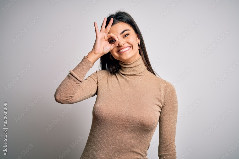 Young beautiful brunette woman wearing turtleneck sweater over white background doing ok gesture with hand smiling, eye looking through fingers with happy face.