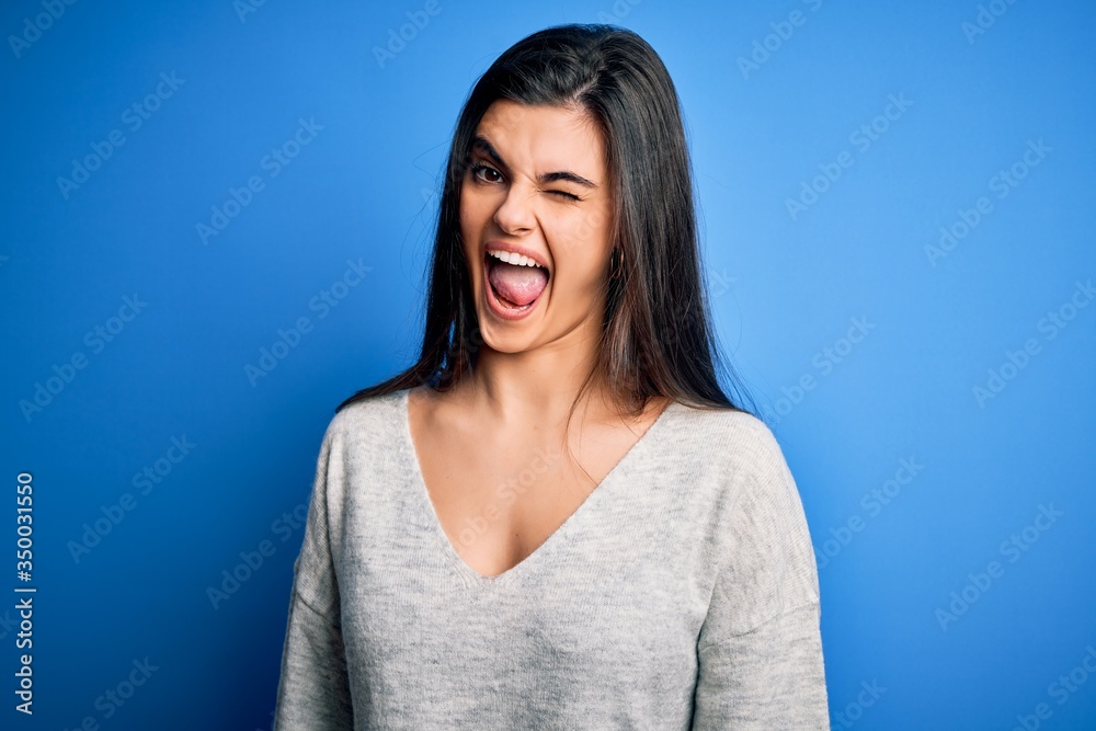 Fototapeta premium Young beautiful brunette woman wearing casual sweater standing over blue background winking looking at the camera with sexy expression, cheerful and happy face.