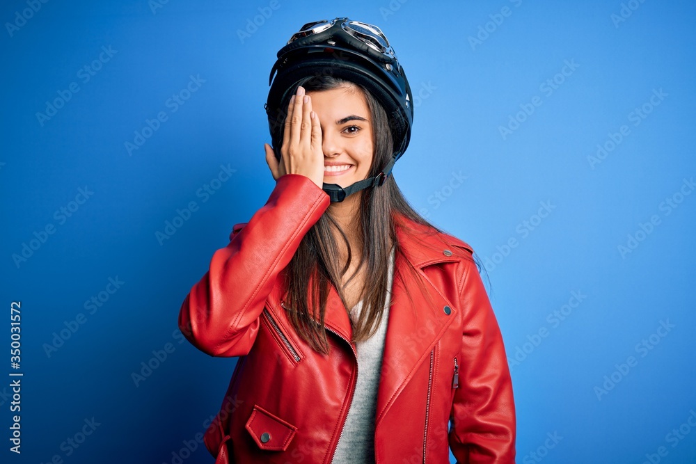 Φωτογραφία Young beautiful brunette motorcycliste woman wearing motorcycle helmet and jacket covering one eye with hand, confident smile on face and surprise emotion