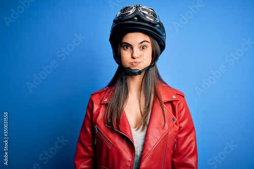 Young beautiful brunette motorcycliste woman wearing motorcycle helmet and jacket puffing cheeks with funny face. Mouth inflated with air, crazy expression. © Krakenimages.com