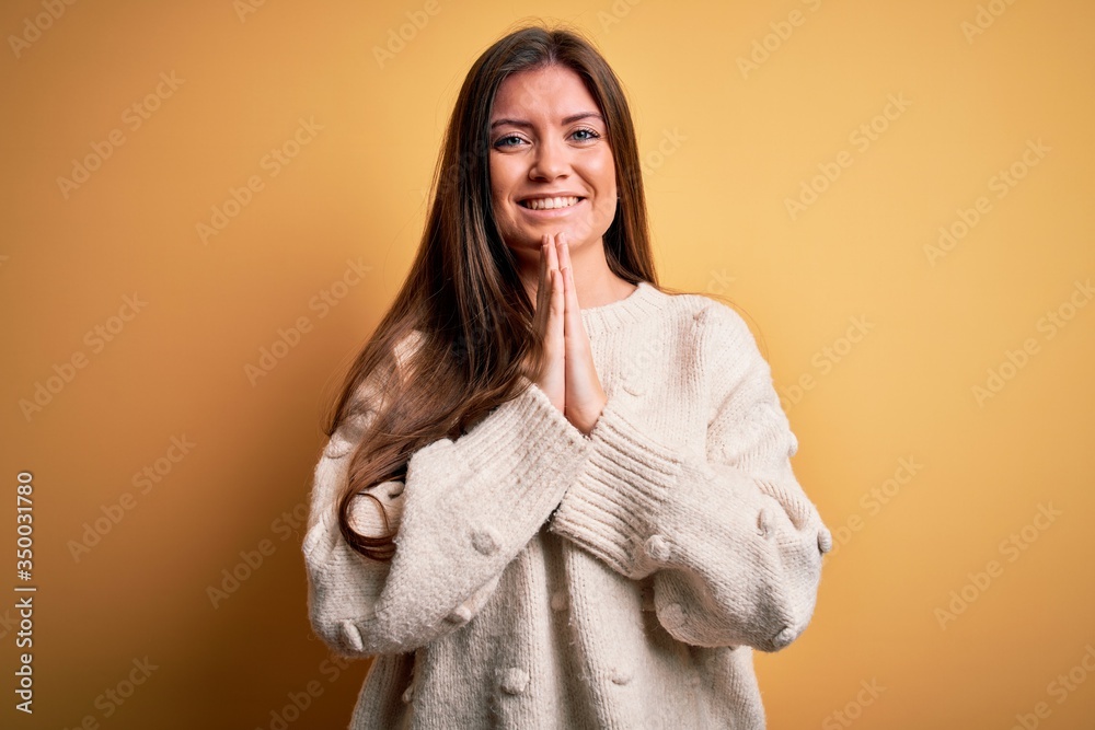 Young beautiful woman with blue eyes wearing casual sweater standing over yellow background praying with hands together asking for forgiveness smiling confident. Young beautiful woman with blue eyes wearing casual sweater standing over yellow background praying with hands together asking for forgiveness smiling confident.