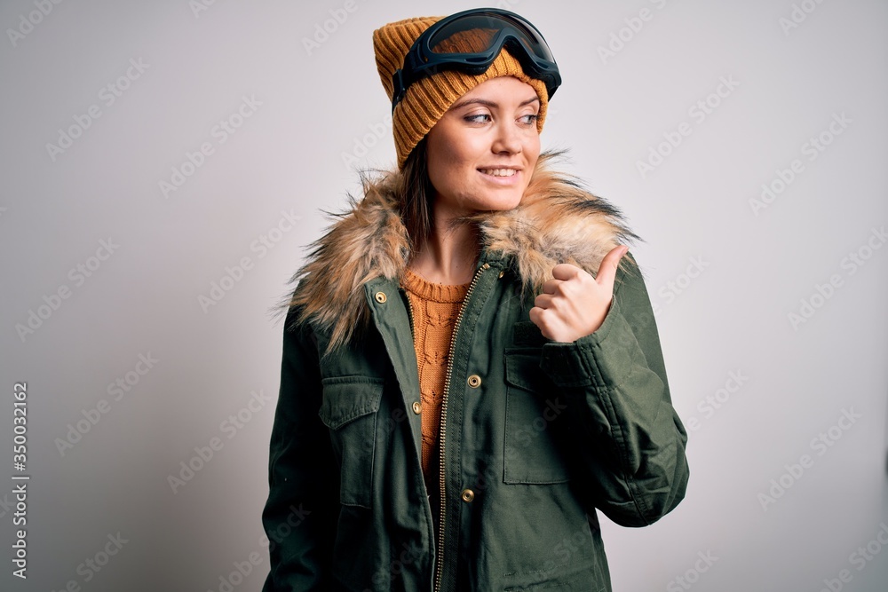 Young beautiful skier woman with blue eyes wearing snow sportswear and ski goggles smiling with happy face looking and pointing to the side with thumb up.