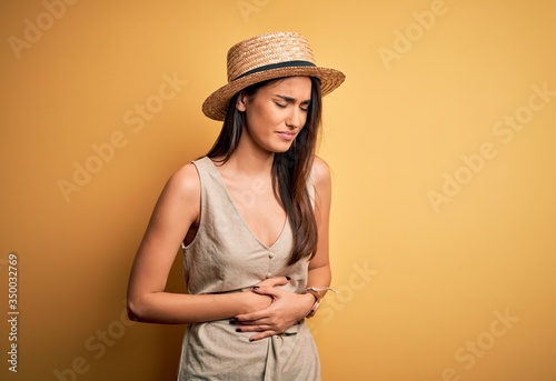 Young beautiful brunette woman on vacation wearing casual dress and hat with hand on stomach because nausea, painful disease feeling unwell. Ache concept.