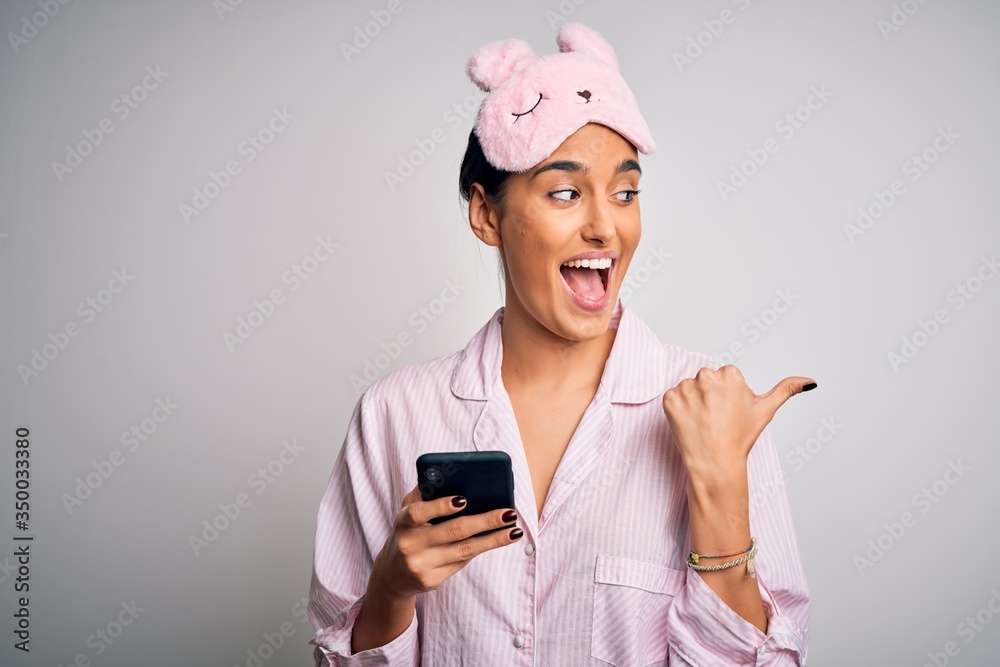 Young beautiful woman wearing pajama and sleep mask having conversation using smartphone pointing and showing with thumb up to the side with happy face smiling Young beautiful woman wearing pajama and sleep mask having conversation using smartphone pointing and showing with thumb up to the side with happy face smiling