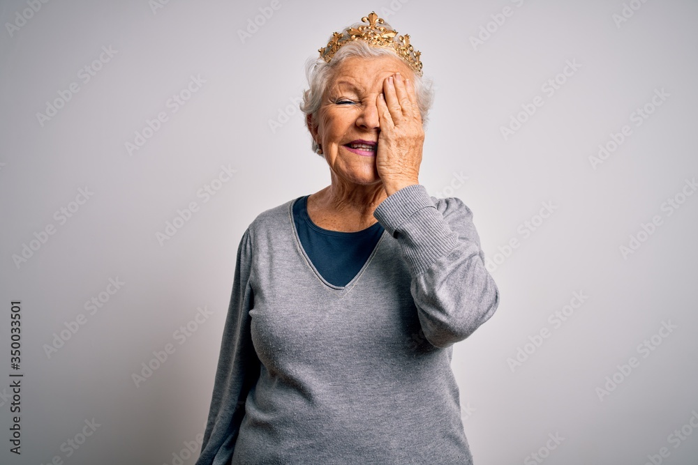 Senior beautiful grey-haired woman wearing golden queen crown over white background covering one eye with hand, confident smile on face and surprise emotion. Senior beautiful grey-haired woman wearing golden queen crown over white background covering one eye with hand, confident smile on face and surprise emotion.
