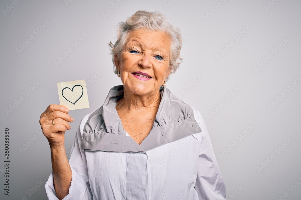 Senior beautiful grey-haired woman holding reminder paper heart over white background with a happy face standing and smiling with a confident smile showing teeth Senior beautiful grey-haired woman holding reminder paper heart over white background with a happy face standing and smiling with a confident smile showing teeth