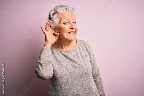 Senior beautiful woman wearing casual t-shirt standing over isolated pink background smiling with hand over ear listening an hearing to rumor or gossip. Deafness concept.