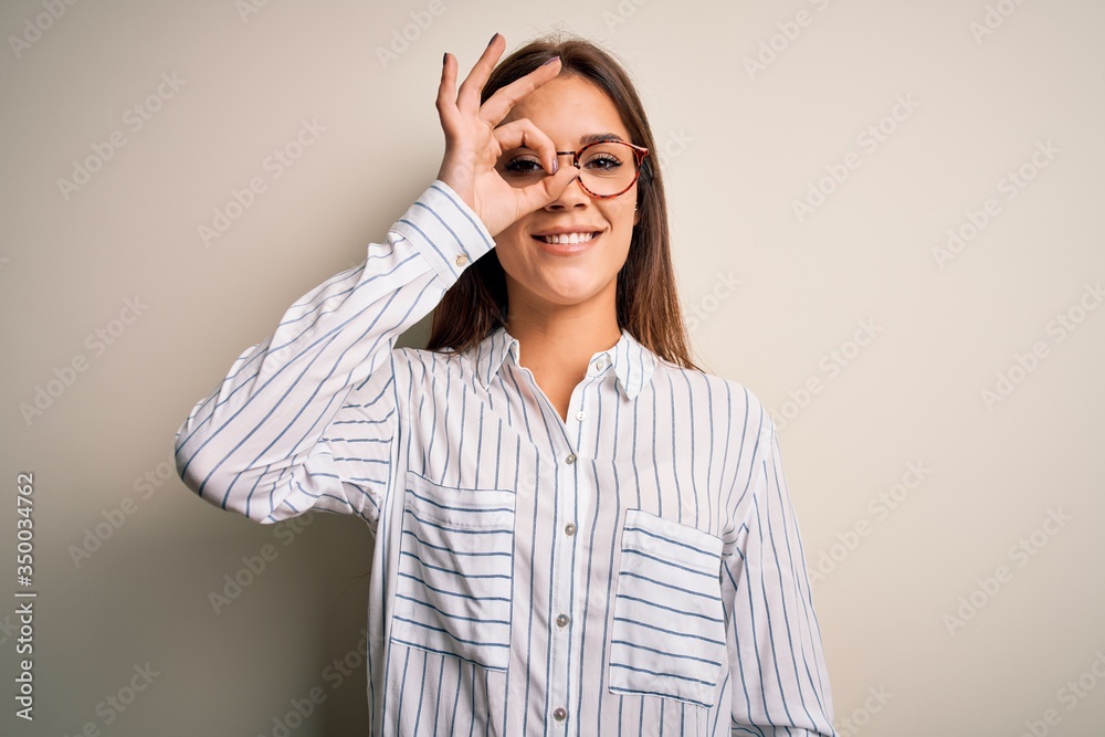Young beautiful brunette woman wearing casual shirt and glasses over white background doing ok gesture with hand smiling, eye looking through fingers with happy face.
