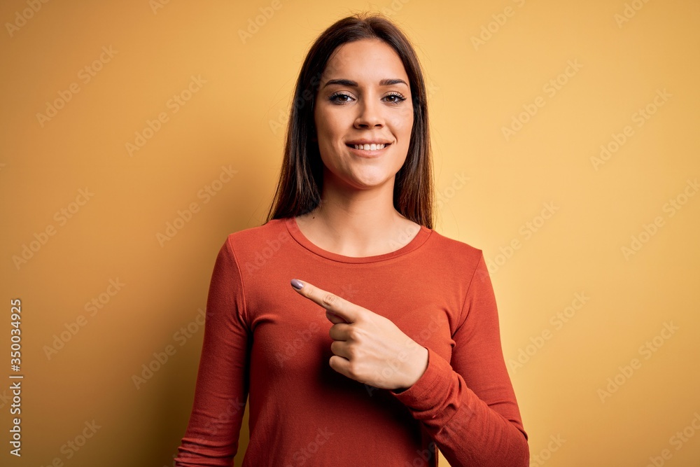 Obraz premium Young beautiful brunette woman wearing casual t-shirt standing over yellow background cheerful with a smile on face pointing with hand and finger up to the side with happy and natural expression