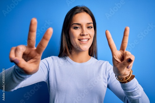 Young beautiful brunette woman wearing casual sweater standing over blue background smiling with tongue out showing fingers of both hands doing victory sign. Number two. © Krakenimages.com