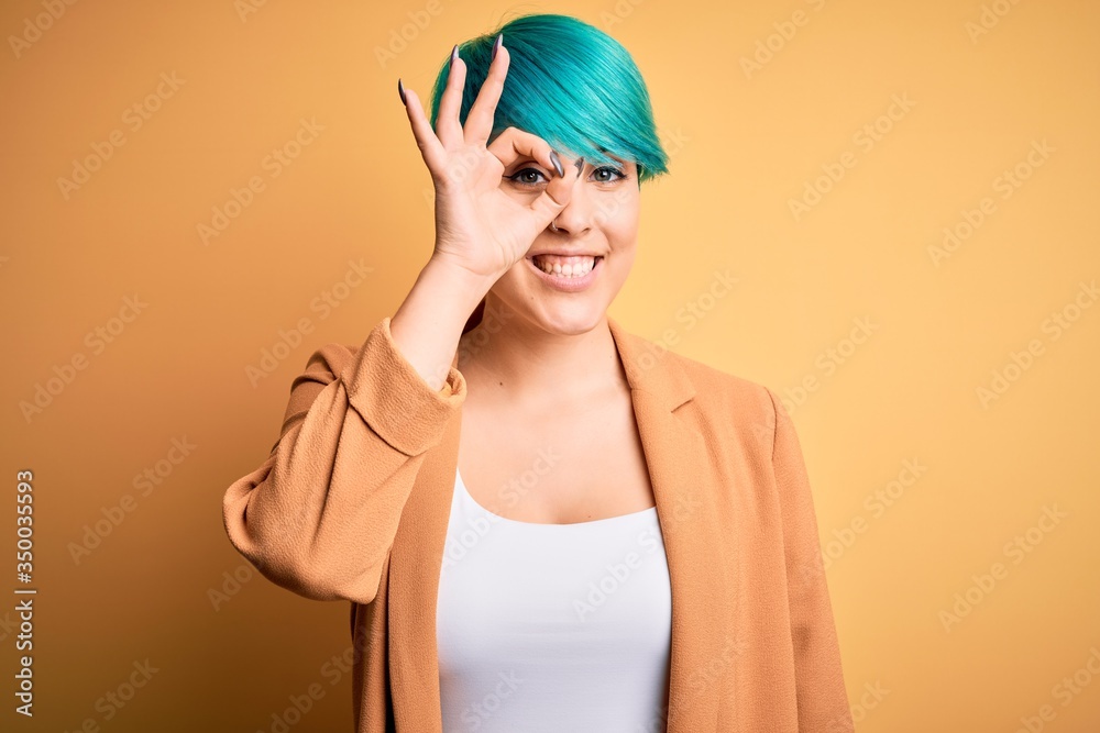 Young beautiful woman with blue fashion hair wearing casual jacket over yellow background doing ok gesture with hand smiling, eye looking through fingers with happy face. Young beautiful woman with blue fashion hair wearing casual jacket over yellow background doing ok gesture with hand smiling, eye looking through fingers with happy face.