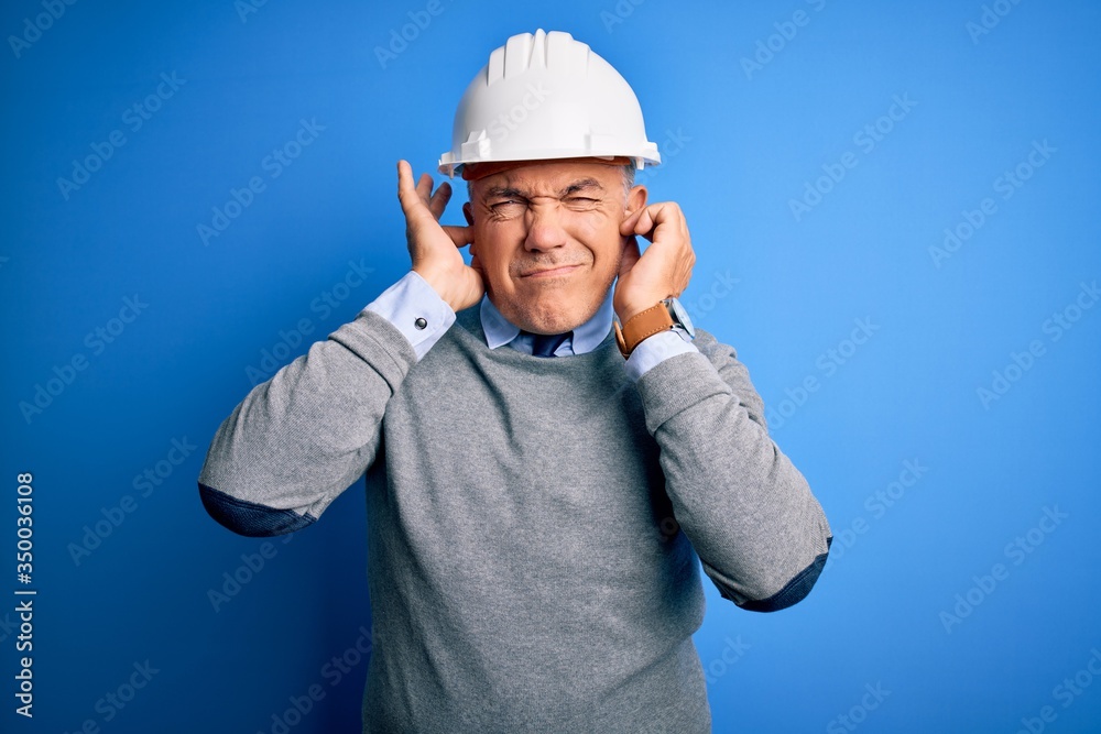 Middle age handsome grey-haired engineer man wearing safety helmet over blue background covering ears with fingers with annoyed expression for the noise of loud music. Deaf concept. Middle age handsome grey-haired engineer man wearing safety helmet over blue background covering ears with fingers with annoyed expression for the noise of loud music. Deaf concept.