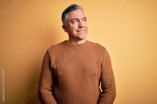 Middle age handsome grey-haired man wearing casual sweater over yellow background looking away to side with smile on face, natural expression. Laughing confident.