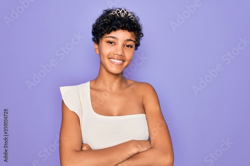 Young beautiful african american afro woman wearing tiara crown over purple background happy face smiling with crossed arms looking at the camera. Positive person.