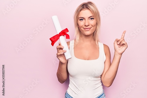Young beautiful blonde student woman holding graduated degree diploma over pink background smiling happy pointing with hand and finger to the side