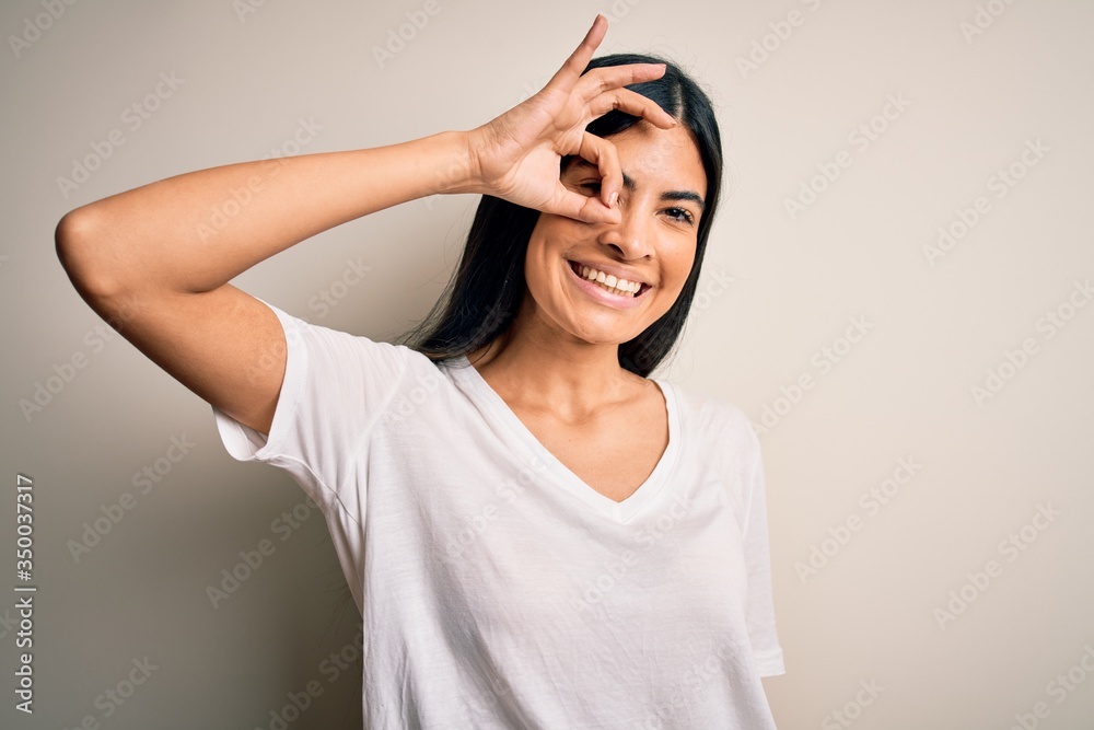 Young beautiful hispanic woman wearing casual white t-shirt over isolated background doing ok gesture with hand smiling, eye looking through fingers with happy face. Young beautiful hispanic woman wearing casual white t-shirt over isolated background doing ok gesture with hand smiling, eye looking through fingers with happy face.