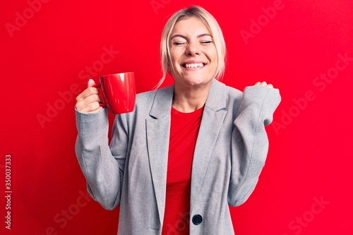 Young beautiful blonde businesswoman drinking cup of coffee over isolated red background screaming proud, celebrating victory and success very excited with raised arm © Krakenimages.com