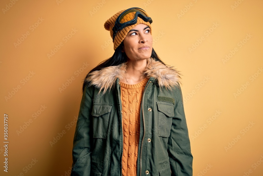 Young beautiful hispanic woman wearing ski glasses and coat for winter weather smiling looking to the side and staring away thinking. Young beautiful hispanic woman wearing ski glasses and coat for winter weather smiling looking to the side and staring away thinking.