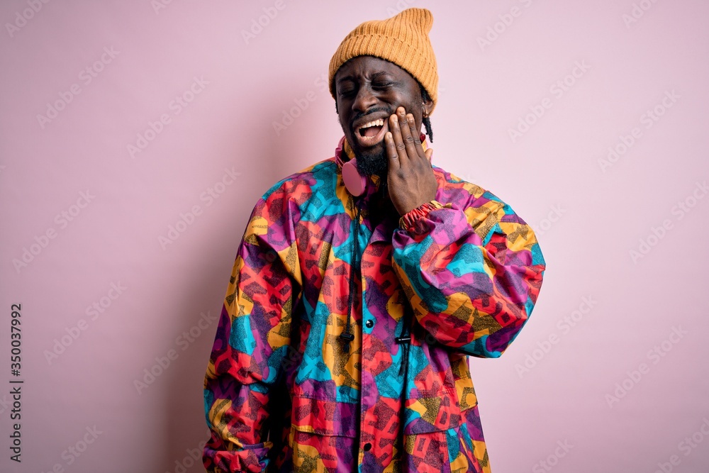 Young handsome african american man wearing colorful coat and cap over pink background touching mouth with hand with painful expression because of toothache or dental illness on teeth. Dentist