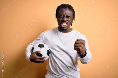Young african american player man playing soccer holding football ball over yellow background annoyed and frustrated shouting with anger, crazy and yelling with raised hand, anger concept