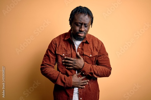 Young handsome african american man wearing casual jacket standing over yellow background with hand on stomach because indigestion, painful illness feeling unwell. Ache concept.
