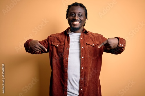 Young handsome african american man wearing casual jacket standing over yellow background looking confident with smile on face, pointing oneself with fingers proud and happy.
