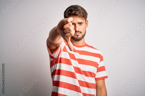 Young handsome man with beard wearing striped t-shirt standing over white background looking unhappy and angry showing rejection and negative with thumbs down gesture. Bad expression.
