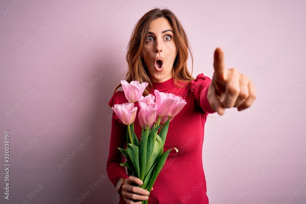 Young beautiful brunette woman holding bouquet of pink tulips over isolated background Pointing with finger surprised ahead, open mouth amazed expression, something on the front Young beautiful brunette woman holding bouquet of pink tulips over isolated background Pointing with finger surprised ahead, open mouth amazed expression, something on the front