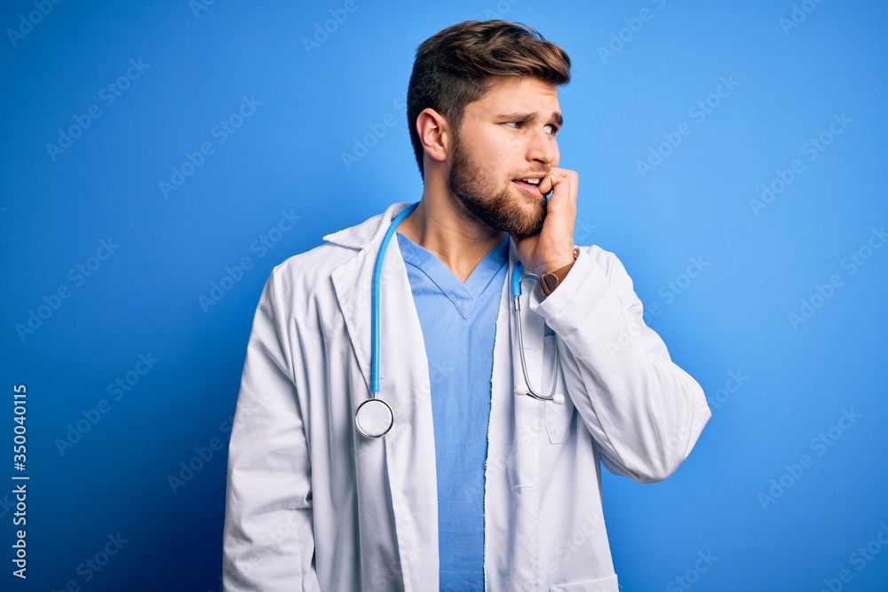 Young blond doctor man with beard and blue eyes wearing white coat and stethoscope looking stressed and nervous with hands on mouth biting nails. Anxiety problem. Young blond doctor man with beard and blue eyes wearing white coat and stethoscope looking stressed and nervous with hands on mouth biting nails. Anxiety problem.