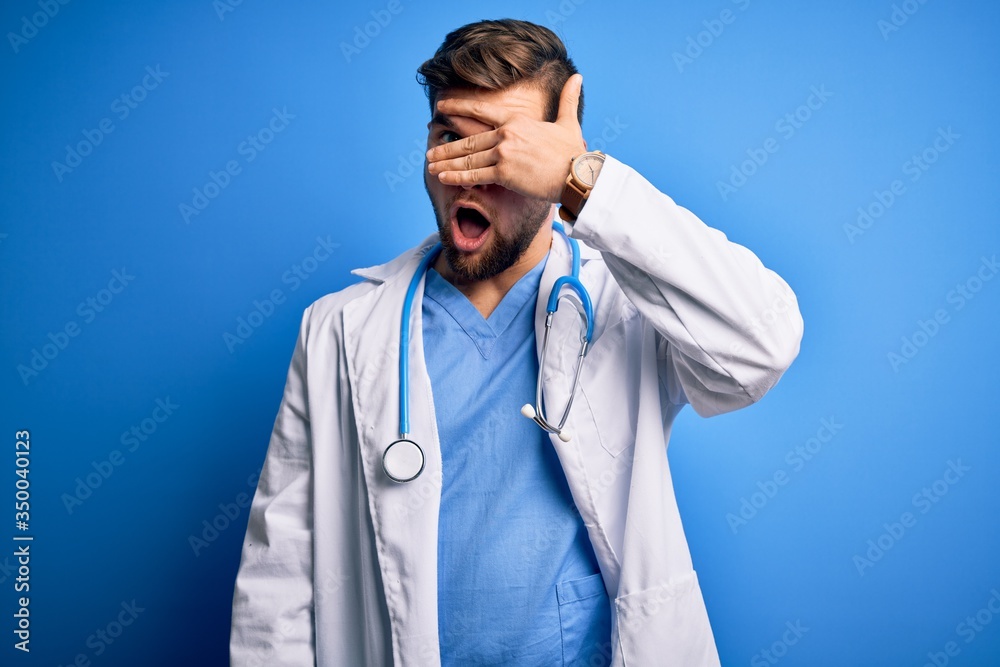 Young blond doctor man with beard and blue eyes wearing white coat and stethoscope peeking in shock covering face and eyes with hand, looking through fingers with embarrassed expression. Young blond doctor man with beard and blue eyes wearing white coat and stethoscope peeking in shock covering face and eyes with hand, looking through fingers with embarrassed expression.