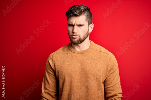Young blond man with beard and blue eyes wearing casual shirt over red background skeptic and nervous, frowning upset because of problem. Negative person.