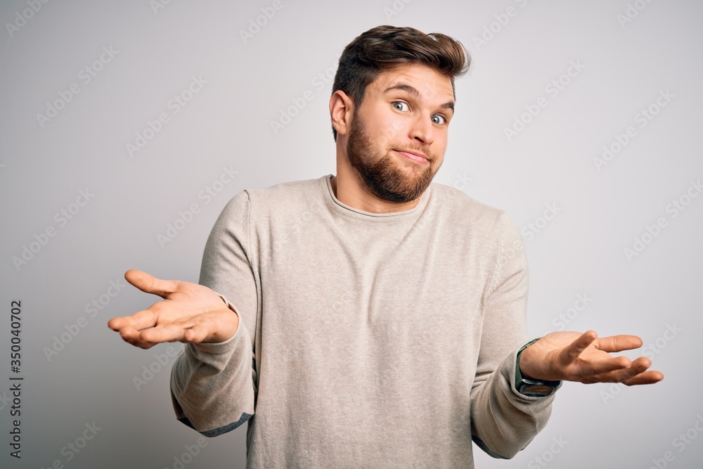 Young handsome blond man with beard and blue eyes wearing casual sweater clueless and confused expression with arms and hands raised. Doubt concept. Young handsome blond man with beard and blue eyes wearing casual sweater clueless and confused expression with arms and hands raised. Doubt concept.