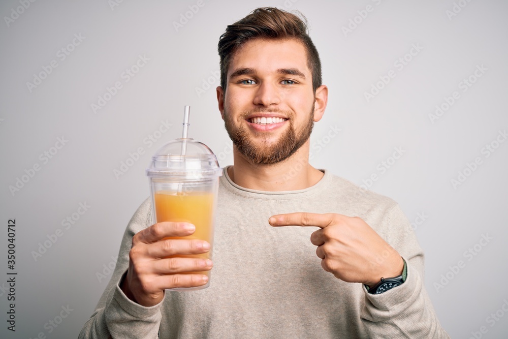 Young blond man with beard and blue eyes drinking healthy orange smoothie very happy pointing with hand and finger Young blond man with beard and blue eyes drinking healthy orange smoothie very happy pointing with hand and finger