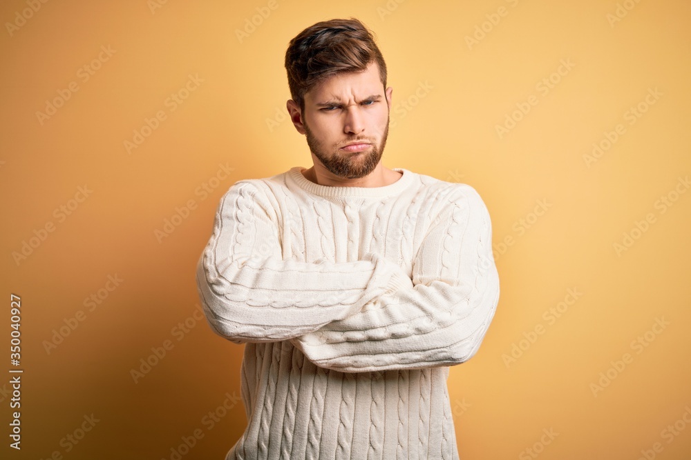 Young blond man with beard and blue eyes wearing white sweater over yellow background skeptic and nervous, disapproving expression on face with crossed arms. Negative person. Young blond man with beard and blue eyes wearing white sweater over yellow background skeptic and nervous, disapproving expression on face with crossed arms. Negative person.