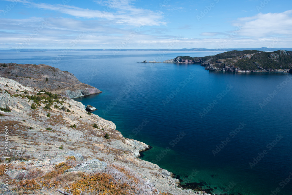 A large cove with deep blue ocean and sky. There are white fluffy clouds near the horizon, The cliffs are ragged and jagged rock. In the distance is a mountain covered in trees and a long rockface. 