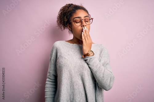 Young beautiful african american girl wearing sweater and glasses over pink background bored yawning tired covering mouth with hand. Restless and sleepiness.